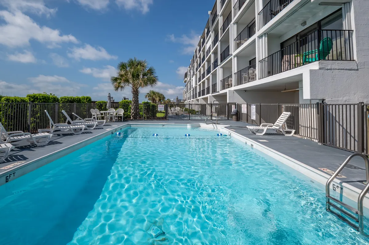 The oceanfront swimming pool is surrounded by palm trees and features several balconies overlooking the water. The scene is lively and welcoming, with sunbathers, swimmers, and relaxed family groups enjoying the pool area, set against the backdrop of the sparkling ocean.