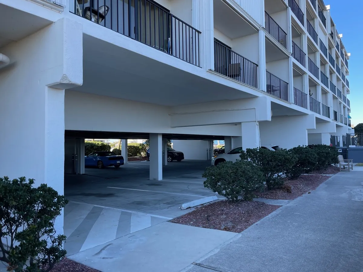 A view underneath the building reveals convenient covered parking, green shrubs lining a pathway, and a casual, laid-back atmosphere. This area is practical for travelers planning day trips and outdoor activities, and the overall scene suggests a friendly community.