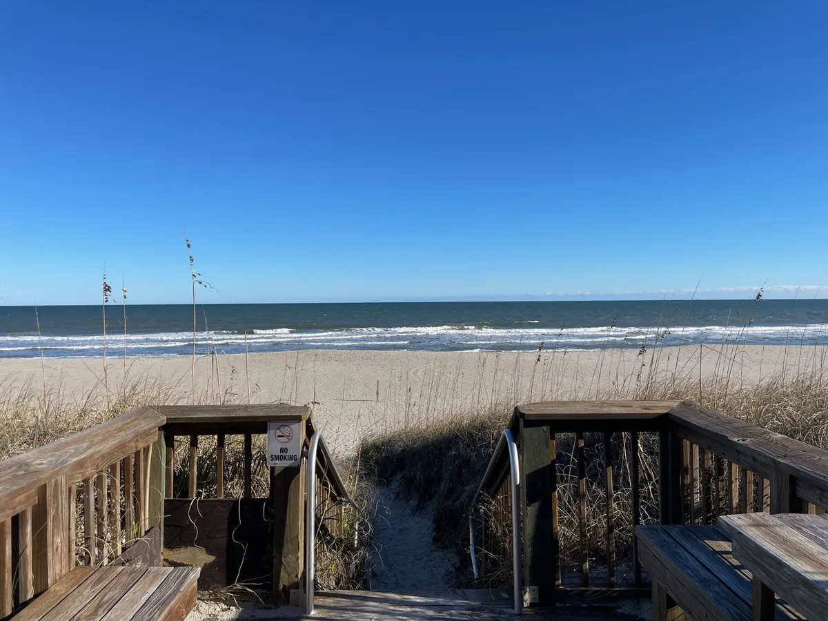 A wooden walkway threads through dune grass and opens directly onto the expansive sandy beach and blue ocean beyond, evoking the sense of arrival at the shoreline. The natural beauty of the dunes and sea is front and center, with families in the distance enjoying classic beach activities.