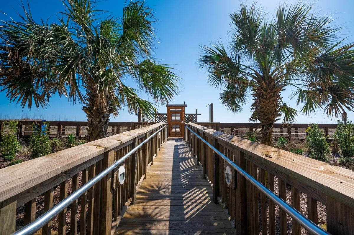 A tranquil wooden boardwalk lined with palm trees that leads to a secure gate at the path’s end, opening to the sandy beach. The scene is peaceful, with hints of ocean breeze and the transition from private property to the beach visible.