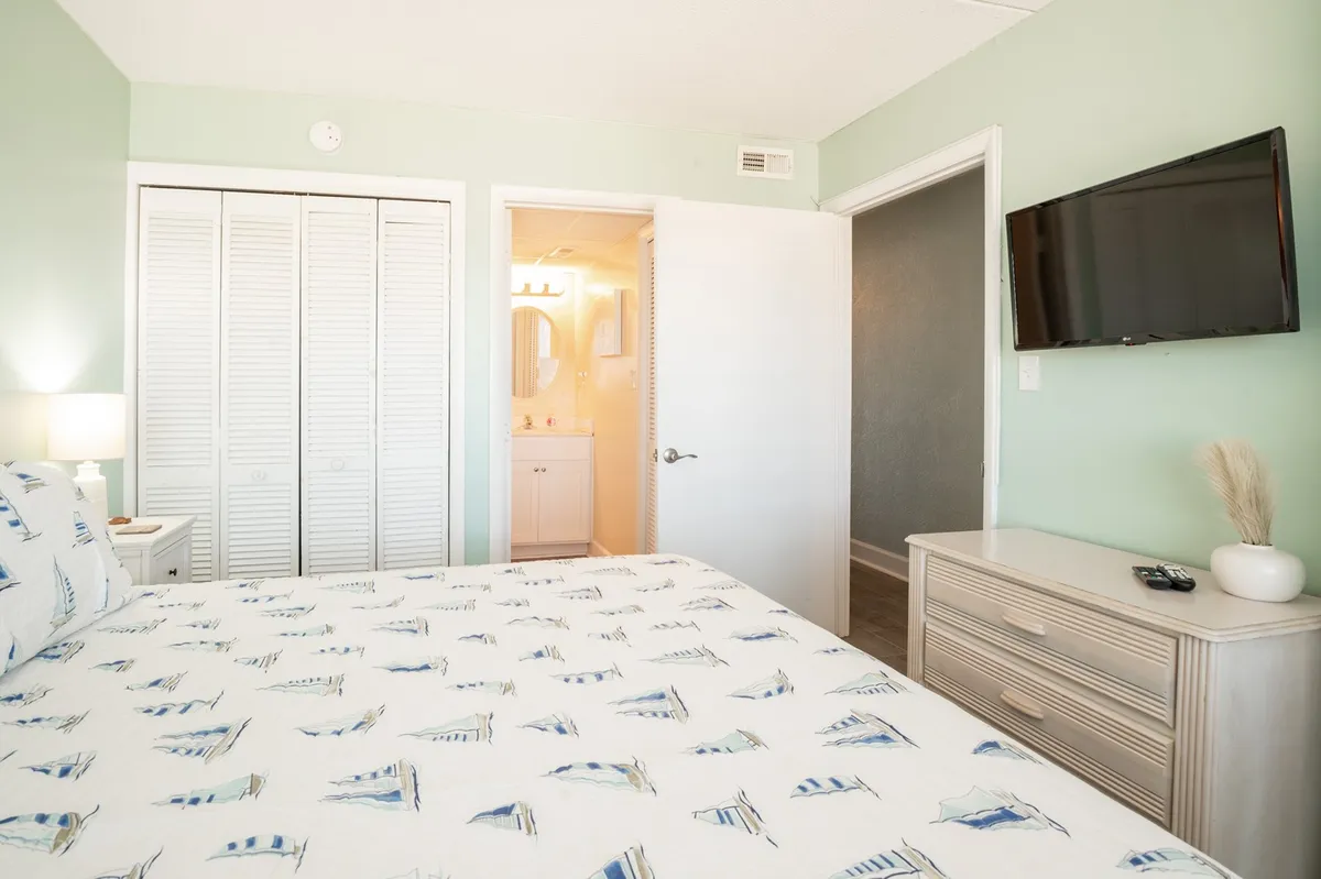 A serene primary bedroom featuring a king-size bed with sailboat-patterned bedding, a wall-mounted smart TV, and a closet with mirrored doors. Soft natural light filters through the window, highlighting the room's clean, comfortable design.