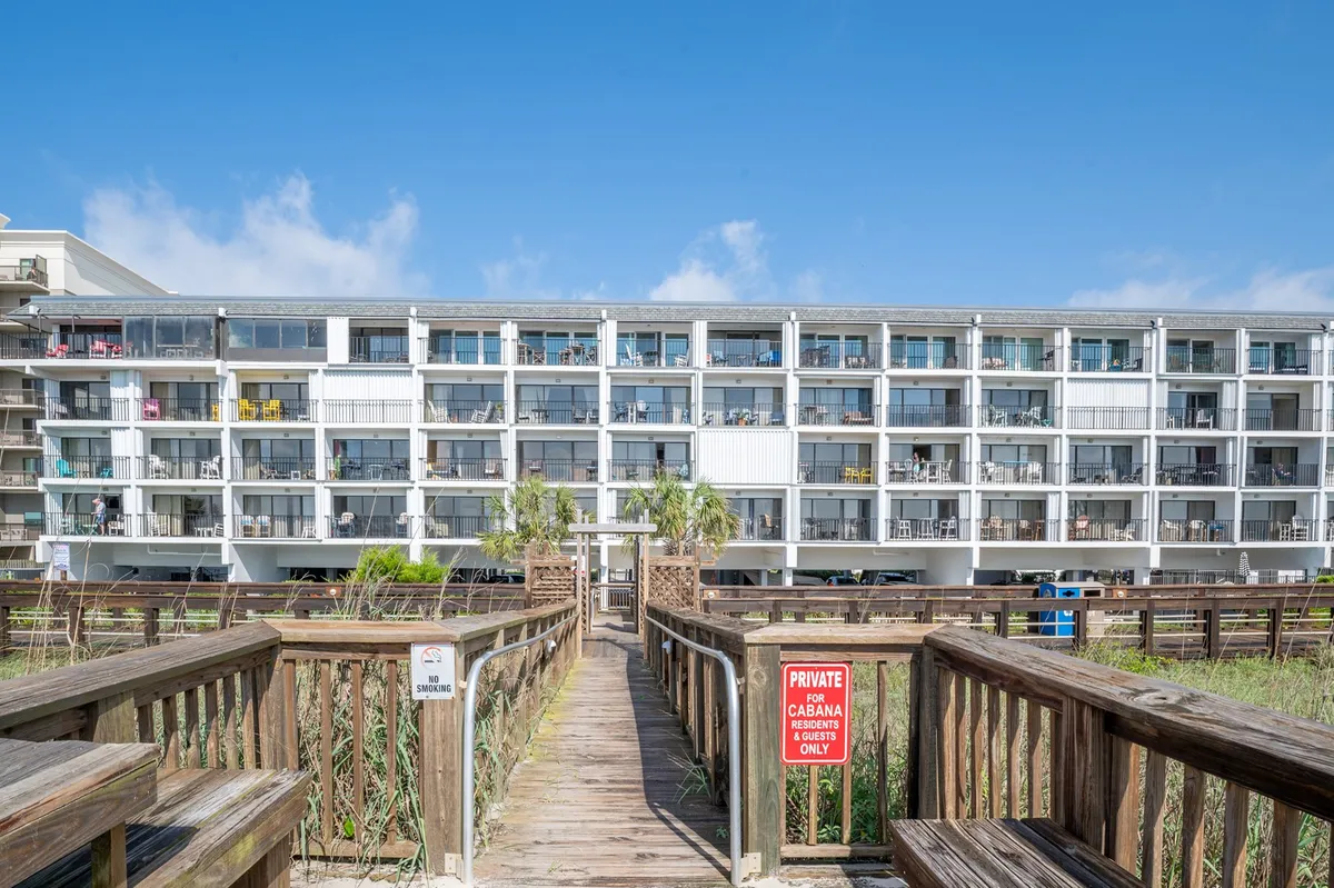 This image captures the exterior boardwalk area by the building facade, with palm trees and signage visible. The pathway leads toward the beach, suggesting easy access to oceanfront views and strolls.