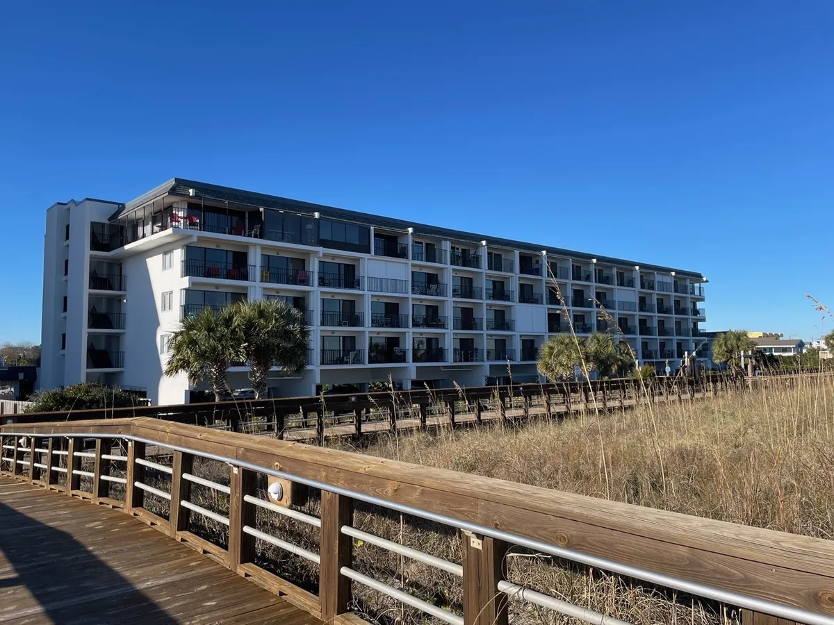 This image captures the modern condominium building set against a backdrop of palm trees, with the Carolina Beach boardwalk visible in the foreground. The scene features people strolling and relaxing, evoking a sense of leisure and connection to the vibrant local area.