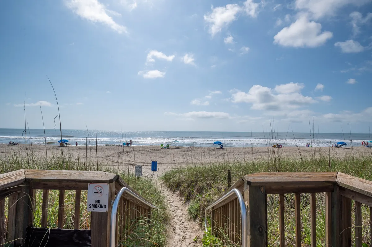 A sweeping exterior view captures the sandy beach, rolling ocean waves, and the private community boardwalk leading guests directly from the property to the shore. Groups of families and children can be seen enjoying beach activities like building sandcastles and beachcombing. The sky is clear and inviting, emphasizing the proximity and ease of access to the beach.