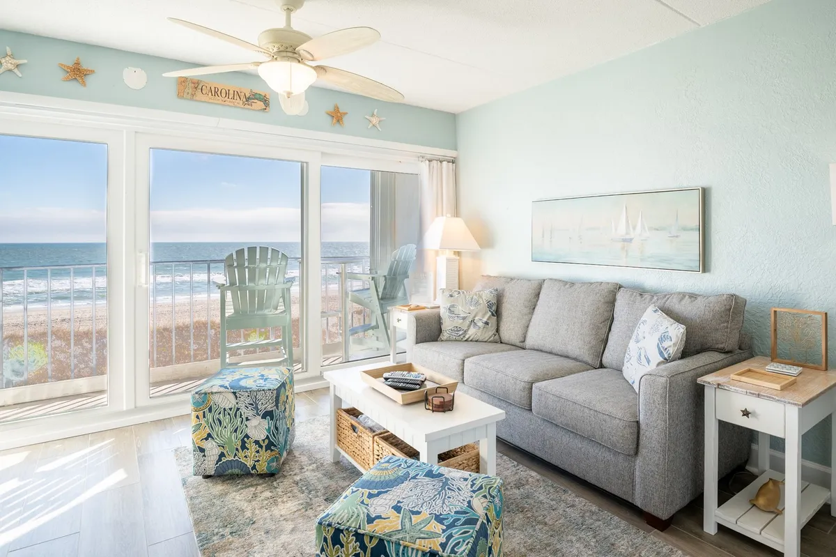 A stylish, sunlit living room with a plush sofa adorned with decorative pillows, a modern coffee table, and an expansive window framing breathtaking ocean views. The space exudes a tranquil, cozy vibe perfect for unwinding, with the Atlantic stretching out just beyond the glass.