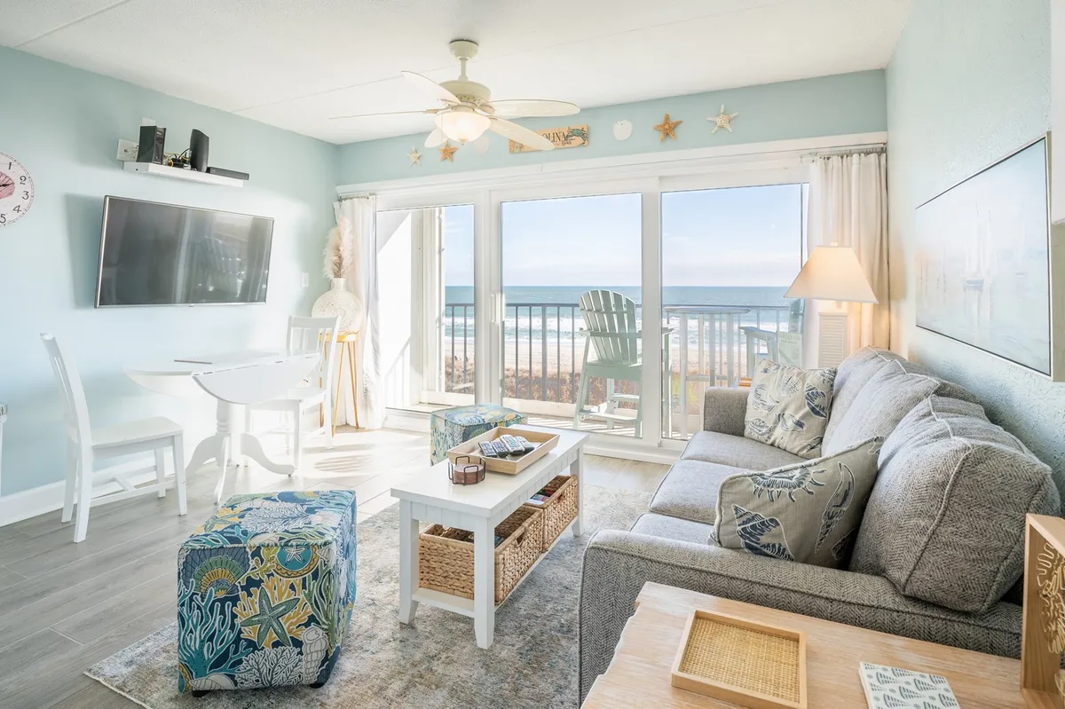 The image showcases a welcoming living area with large windows framing an expansive ocean view. A comfortable sofa with decorative pillows and a stylish coffee table create an inviting atmosphere for relaxation and group gatherings. The space is bright and cheerful, emphasizing both the connection to the outdoors and the comfort of the interior.