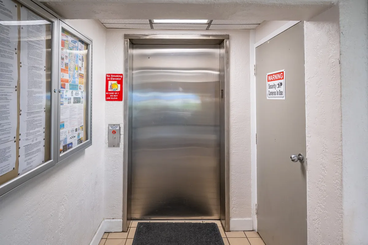An interior shot featuring elevator doors, an informational bulletin board, and warning signs in a well-lit lobby. The space is practical, clean, and welcoming, suggesting efficient elevator service and community communication.